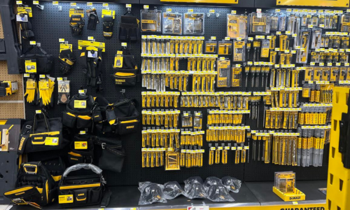 Display of DEWALT hand tools and accessories, including gloves, tool belts, measuring tapes, drill bits, and utility knives, all neatly organized on a store wall.